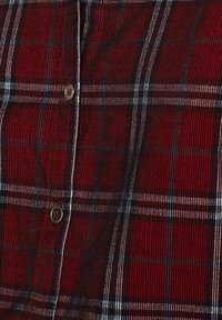 Camicia a quadri rossa e nera con strisce bianche, dotata di un tessuto testurizzato e chiusura frontale con bottoni. Accentuata da bottoni metallici.