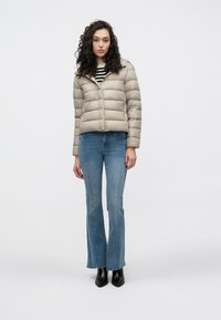 Béžová prešívaná bunda s horizontálnym prešívaním, zapínaním na zips a golierom, kombinovaná s rozšírenými modrými džínsami a čiernymi členkovými čižmami.