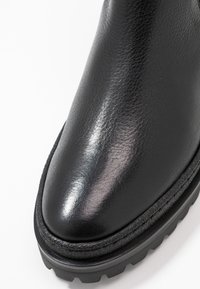 Musta nahkainen nilkkabootsi, jossa on kiiltävä pinta, pyöreä kärki, teksturoitu pohja ja erottuva ommel reunassa.