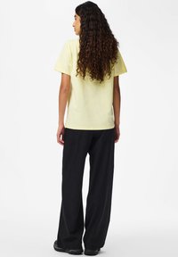 Jasnożółta bawełniana koszulka z krótkim rękawem, zestawiona z czarnymi spodniami w cienkie paski. Widok od tyłu ukazuje luźny krój i teksturowane włosy.
