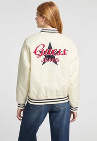 Crème bomberjack met zwarte gestreepte geribbelde manchetten en Zoom. Heeft een groot rood en zwart geborduurd "Guess Jeans" logo met sterdetail op de achterkant.
