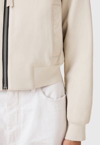 Chaqueta de cuero color crema con un diseño recortado, puños acanalados y una cremallera visible. Incluye bolsillos laterales y una textura suave.