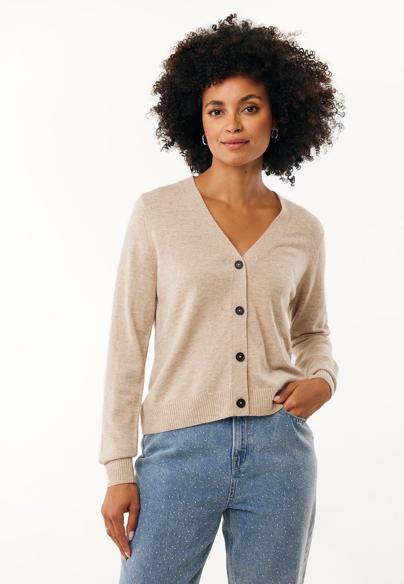 Béžový pletený kardigan s výstřihem do V a černým zapínáním, s žebrovanými manžetami a lemem, nošený přes modré džíny s tečkovaným vzorem.