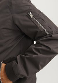 Black bomber jacket featuring a zippered pocket on the left arm, ribbed cuffs, and smooth fabric texture.