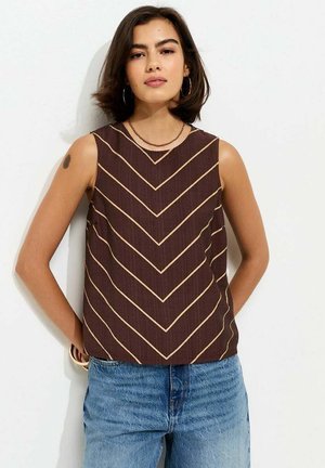 Giovane donna con capelli scuri fino alle spalle che indossa una canottiera marrone con strisce beige a zigzag e jeans blu, in piedi contro un muro bianco.