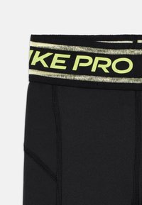 Melnas sporta legingi ar platu jostasvietu, uz kuras ir spilgti dzeltenā krāsā uzraksts "NIKE PRO", un gluda, elastīga auduma virsma ar redzamām šuvju detaļām.