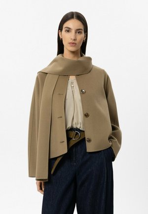 Frau mit langen dunklen Haaren, die eine beige, geknöpfte Jacke mit befestigtem Schal, eine weiße Bluse, dunkle Hose und einen Gürtel mit einem silbernen Ring trägt.