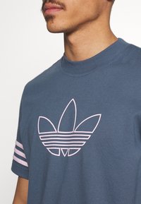 Modrá bavlněná tričko s kulatým výstřihem, které má růžové logo se třemi listy a pruhované akcenty na rukávech. Měkká textura, standardní střih.