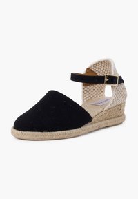 Zwarte canvas wedge espadrille met een geweven witte enkelband en een jute touwzool. Beschikt over een ronde neus en een verstelbare band.