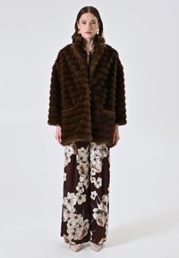 Giacca in pelliccia marrone con righe verticali, caratterizzata da un collo sciallato e tasche laterali. Indossata sopra pantaloni a palazzo con motivo floreale in colori ricchi.