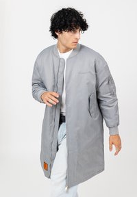 Man met krullend zwart haar, gekleed in een lange grijze bomberjack over een witte shirt en lichtblauwe jeans, kijkend naar de zijkant.