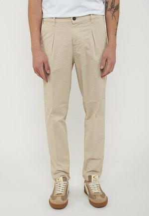Uomo con pantaloni beige plissettati e sneakers beige e bianche in piedi davanti a uno sfondo bianco.