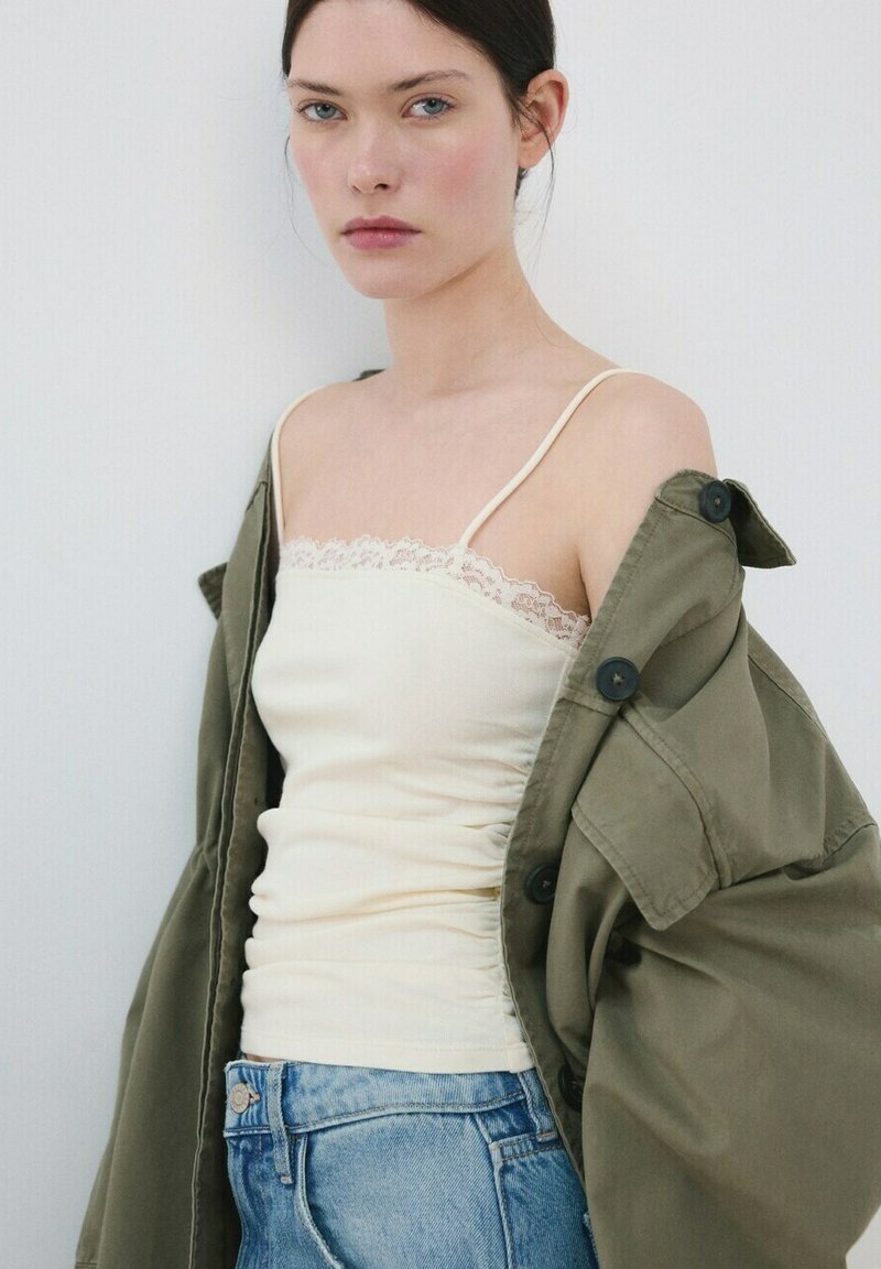 Jeune femme aux cheveux foncés portant un débardeur crème orné de dentelle, une veste ample vert olive et un jean bleu clair sur un fond uni.