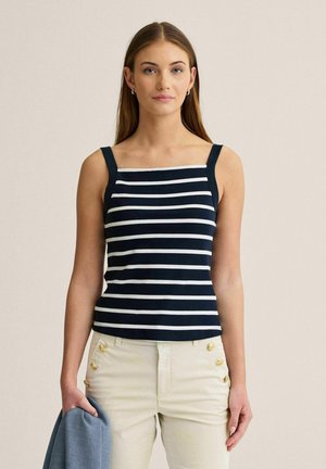 Nuori nainen, jolla on pitkät hiukset ja joka pukeutuu tummansinivalkoraidalliseen hihattomaan toppiin ja kermaan vivahtaviin housuihin, joissa on kultaiset napit. Hän pitää vasemmassa kädessään sinistä kangasta.