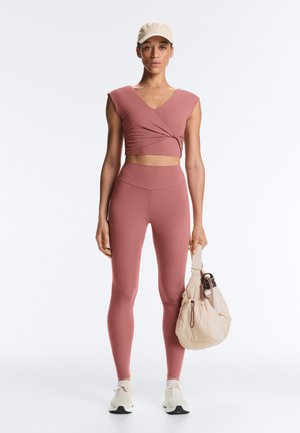 Žena ve světle purpurovém zavinovacím crop topu a vysokým pasem legínách, krémová čepice, bílé tenisky, drží béžovou kabelku na jednobarevném pozadí.