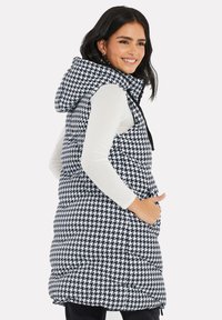 Pufferväst i houndstooth-mönster i svart och vitt, med huva, sidfickor och quiltad textur. Utrustad med hög krage och frontdragkedja.