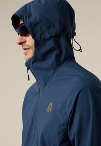 Hombre con chaqueta azul marino con capucha y cremallera, y gafas de sol oscuras, mirando hacia la izquierda, sobre un fondo claro.