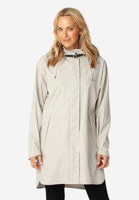 Donna che indossa un impermeabile lungo grigio chiaro con collo alto, fibbia nera, tasche laterali e pantaloni neri, in piedi davanti a uno sfondo semplice.