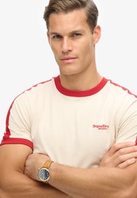 Cremefarvet T-shirt med røde detaljer på kraven og ærmerne, der har "SuperDry Athletics" logo. Modellens håndled prydes af et guldur.