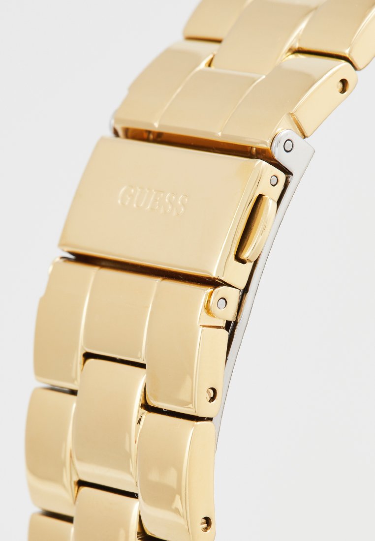 Pulsera de reloj de metal dorado con eslabones rectangulares, acabado pulido y un cierre que presenta el nombre de la marca "GUESS" grabado en él.