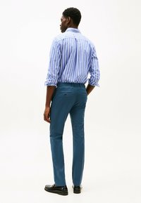 Camicia a righe blu e bianca con maniche arrotolate, abbinata a pantaloni teal e mocassini neri. Focus sulla vestibilità e sulle linee pulite.