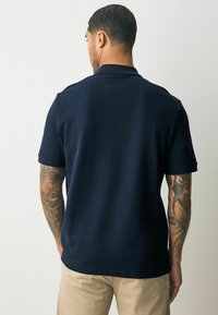 Marine polo skjorte med korte ærmer, tekstureret stof og klassisk krave, parret med brune bukser. Synlig tatovering på armen.