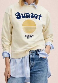 Kremowy sweatshirt z napisem "Sunset" i "Groovy 1976" w ciemnoniebieskim kolorze. Charakteryzuje się żółtym okrągłym wzorem w paski. Noszony na niebieskiej koszuli w paski.