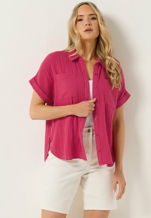 Vrouw met lang blond haar die een magentakleurig overhemd met korte mouwen en knopen draagt over een witte top en crèmekleurige shorts tot aan de knie, staand tegen een lichte achtergrond.