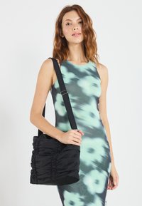 Schwarze, geraffte Handtasche aus Stoff mit glatter Textur, verstellbarem Tragegurt und einer weichen, abgerundeten Form. Getragen über einem blumengemusterten Kleid.