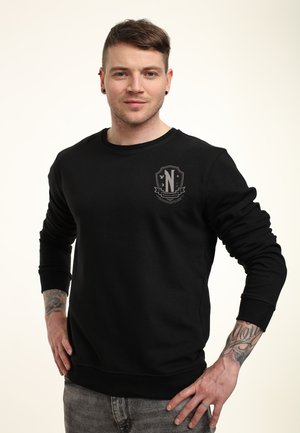 Jonge man met kort bruin haar, gekleed in een zwart sweatshirt met een embleem, staande met de handen op zijn heupen en tattoos op zijn onderarmen.