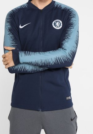 Marineblaue Zip-Jacke mit hellblauen, strukturierten Ärmeln, Chelsea FC-Logo auf der Brust, Nike-Swoosh, getragen von einer Person mit verschränkten Armen.
