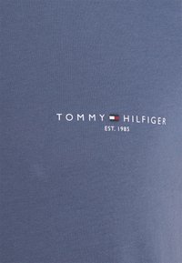 Tissu bleu avec le texte blanc "Tommy Hilfiger Est. 1985" et un petit logo rectangulaire avec des couleurs rouge, blanc et bleu centré entre les mots.