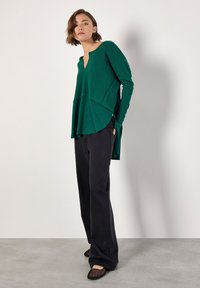 Grünes, geripptes Oberteil mit V-Ausschnitt und geschichtetem Saum; kombiniert mit schwarzen, weiten Hosen und flachen Schuhen. Einfaches Design mit strukturierten Stoffakzenten.