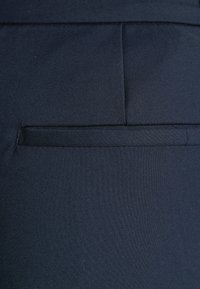 Jūras spēku zila auduma ar gludu tekstūru, kam ir diagonāla kabatas detaļa. Materiāls ir viegls un austs, ar minimāli redzamu šuvju.