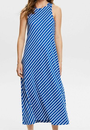 Femme portant une robe bleue sans manches avec des rayures blanches diagonales et un col rond, debout devant un fond clair uni.