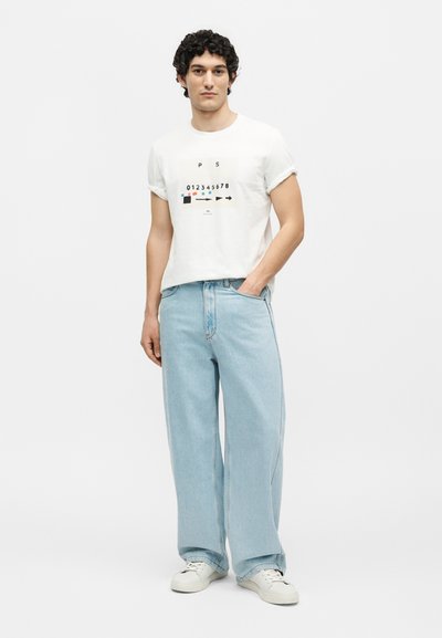 Νέος με σγουρά μαλλιά που φοράει λευκό γραφικό μπλουζάκι, ανοιχτό μπλε τζιν παντελόνι με φαρδιά πόδια και λευκά αθλητικά παπούτσια, στέκεται μπροστά σε ένα απλό φόντο.