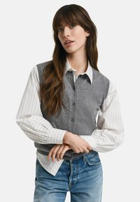 Gilet grigio sopra una camicia bianca a righe con bottoni, caratterizzata da un fit rilassato, dettagli con bottoni e maniche lunghe a sbuffo. I jeans blu completano il look.