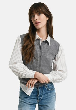 Gray vest over a white and striped button-up shirt, featuring a relaxed fit, button details, and long, gathered sleeves. Blue jeans complete the look.