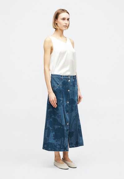 Γυναίκα με κοντά ξανθά μαλλιά που φοράει λευκό αμάνικο τοπ, μπλε τζιν φούστα μέσης με κουμπιά και μπεζ φλατ παπούτσια, στέκεται μπροστά σε απλό φόντο.