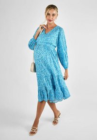 Blaues, gemustertes Kleid mit V-Ausschnitt, langen voluminösen Ärmeln und ausgestellt geschnittenem Saum, kombiniert mit beigen Riemchensandalen und einer kleinen Tasche.