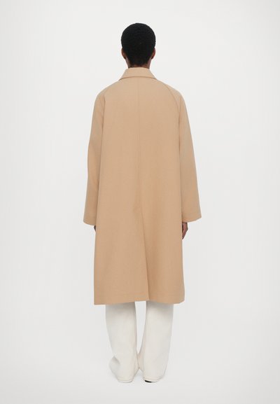 Beige wool coat with a straight cut, large collar, and dropped shoulders. Features a clean back design and is paired with white wide-leg pants.