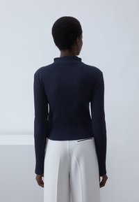 Top de manga larga acanalado en color navy con cuello alto, diseño ajustado y un detalle de costura visible en la parte posterior; combinado con pantalones blancos de corte entallado.