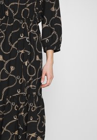 Svart klänning med ett mönster av kedjor och fransar i guld, med långa puffärmar och en insvängd midja som framhäver en svävande silhuett.