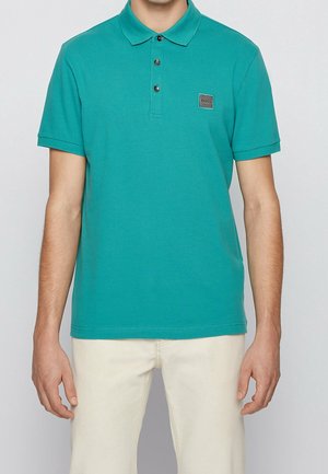 Mand iført en turkis kortærmet polo med krave og tre knapper, kombineret med lyse beige bukser.