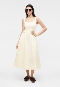 Femme portant une robe midi sans manches écrue, lunettes de soleil foncées et chaussures marron à pampilles, debout avec la main sur la taille.