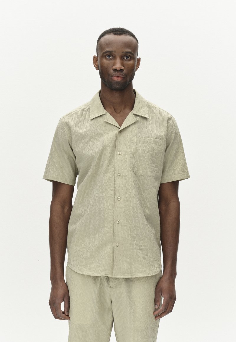 Camicia a maniche corte color oliva con un motivo testurizzato, abbottonata frontalmente e con una tasca sul petto. Indossata con pantaloni abbinati.
