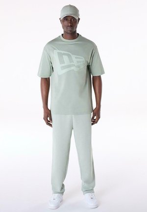 Man wearing light green matching cap, oversized t-shirt with logo, sweatpants, and white sneakers, standing against white background.