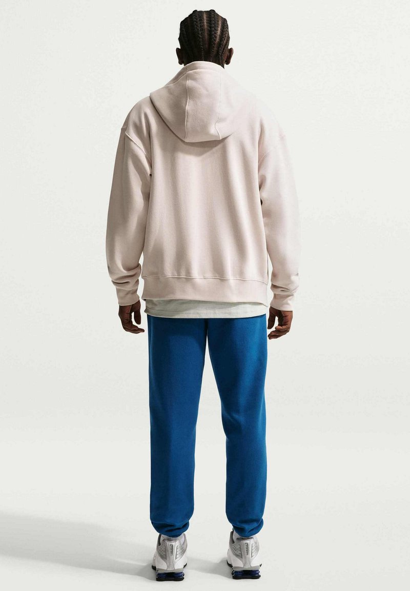 Ljusrosa luvtröja med avslappnad passform. Blå sweatpants med avsmalnande ben. Vita sneakers med texturerade sulor. Enkelt design utan synliga logotyper.