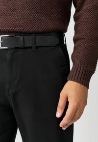 Fekete nadrág sötétbarna bordázott pulóverrel párosítva. Egy fekete bőrszíj látható ezüst csattal. A textúrák kontrasztjára helyeződik a hangsúly.