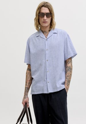 Άνδρας με μακριά μαλλιά και τατουάζ που φοράει γυαλιά ηλίου, ανοιχτού μπλε κοντομάνικο πουκάμισο με κουμπιά, μαύρο παντελόνι, κρατώντας ένα καφέ σακίδιο.